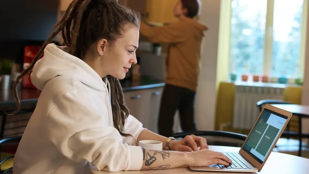Woman in a white hoodie working on a laptop at home with coffee on the table and another person in the background
