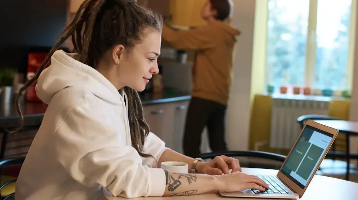 Woman in a white hoodie working on a laptop at home with coffee on the table and another person in the background