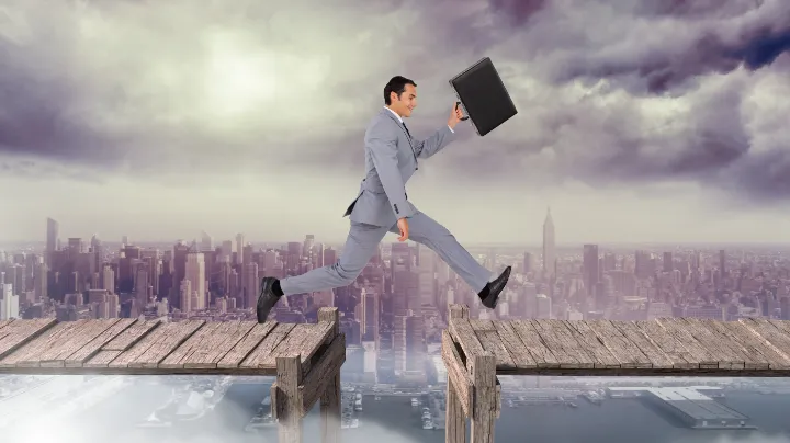 A business professional in a suit leaps over a broken bridge between two platforms high above a city skyline, symbolizing overcoming gaps and challenges in recruitment vendor partnerships.